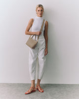 Person standing against wall holding small handbag wearing sleeveless top, pants, sandals.