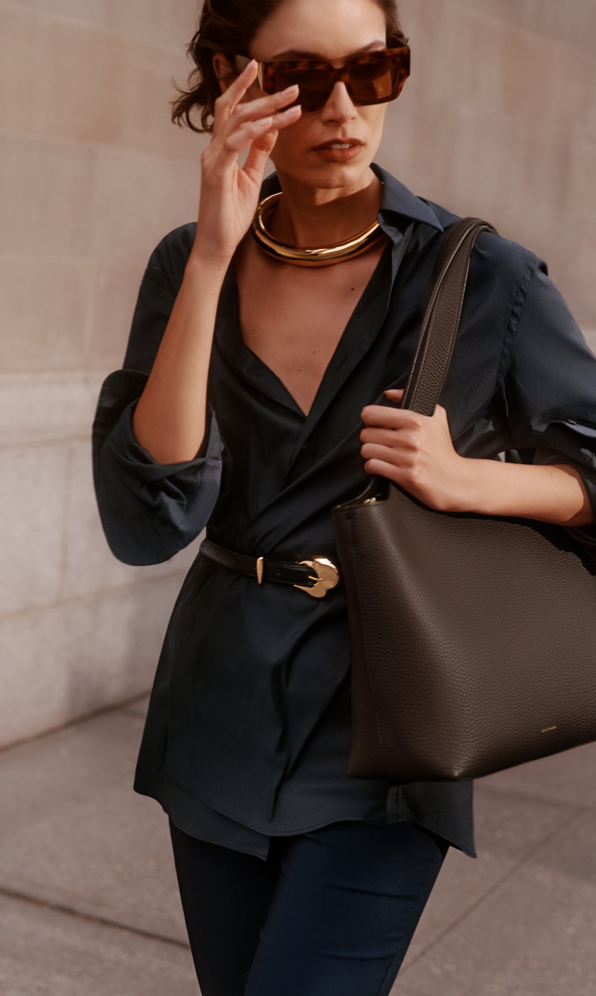 Person wearing sunglasses and dark outfit holding a handbag, with a gold necklace and belt.