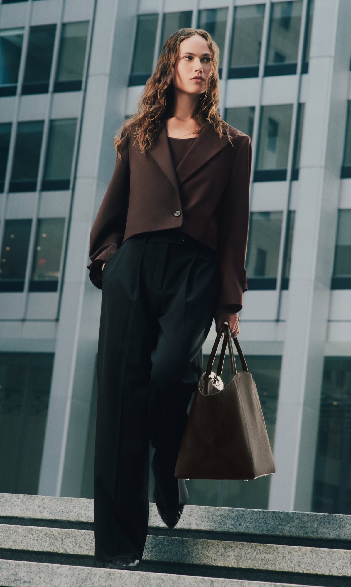 Person in a cropped jacket holding a large bag, standing near tall buildings.