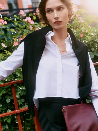 Woman in a white shirt with a sweater draped over her shoulders, holding a handbag.