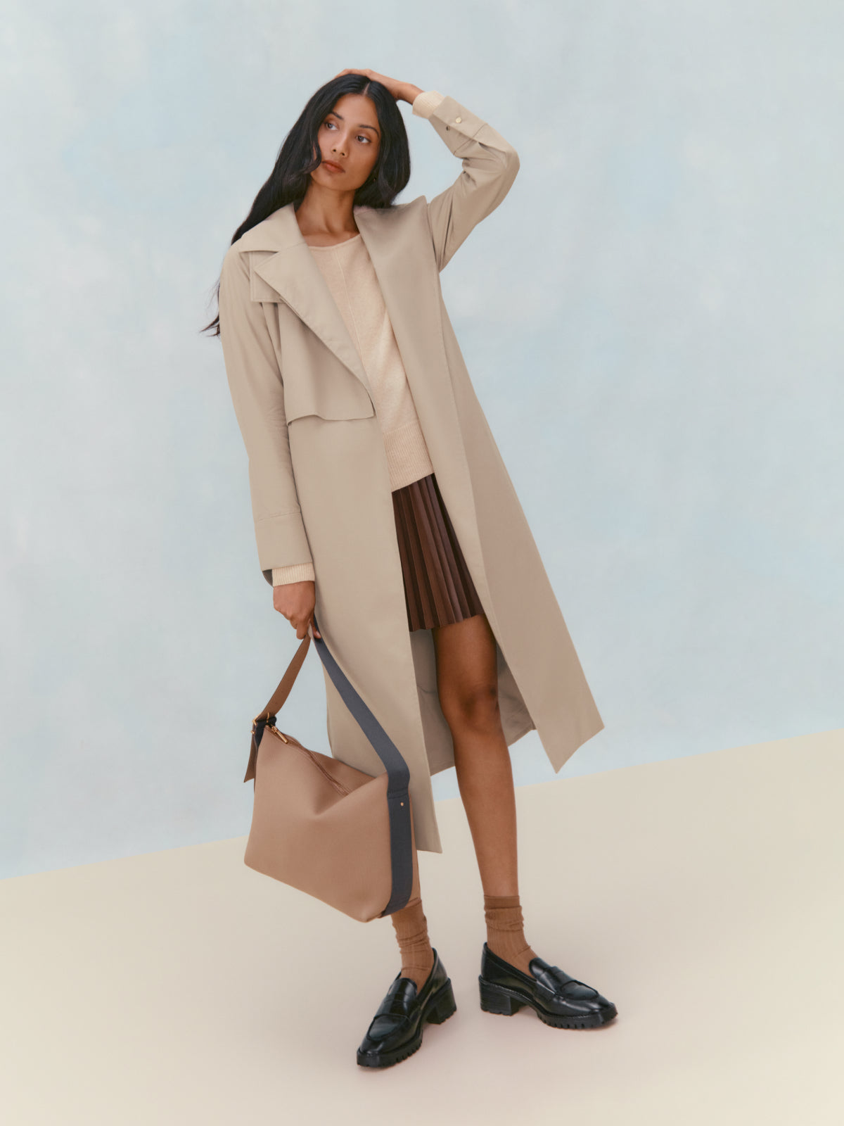Woman standing with hand on head, holding a bag and wearing a long coat and boots.