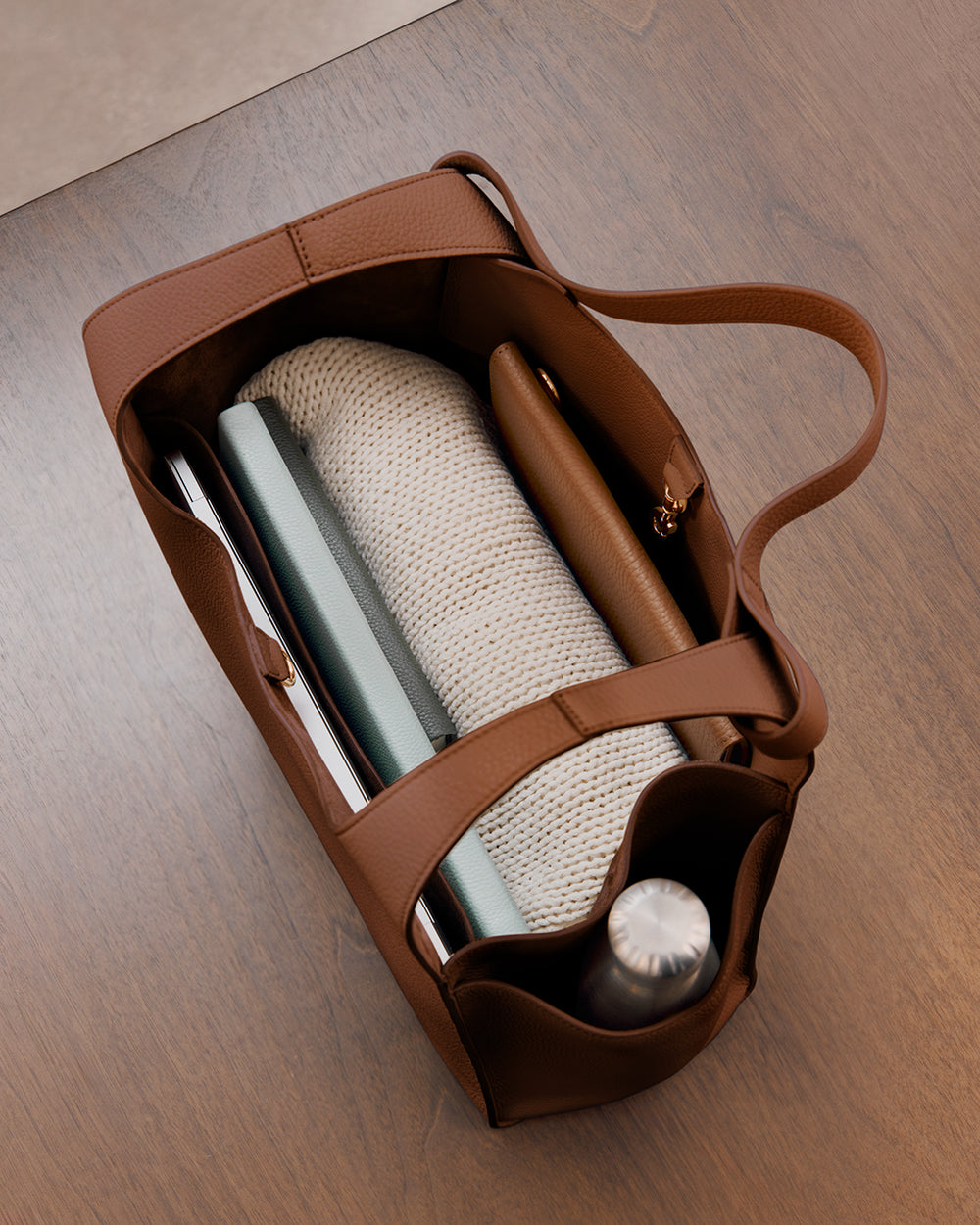 Top view of an open handbag containing a book, a notebook, a rolled-up sweater, a wallet, and a water bottle.