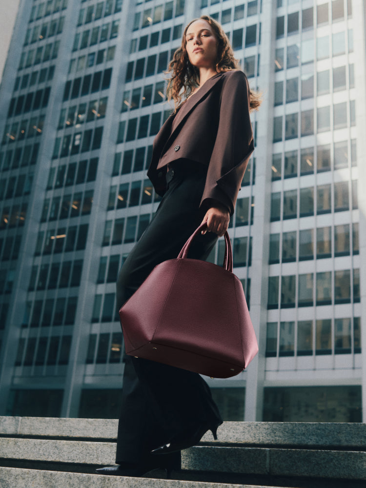 Person stands on steps holding a large handbag in front of tall buildings.