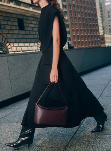 Person walking in urban setting carrying handbag wearing long skirt, asymmetrical top and boots