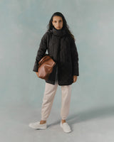 Woman standing with a coat, scarf, pants, holding a bag, and wearing sneakers.