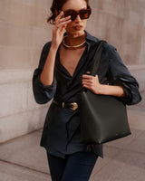 Person wearing sunglasses, holding a bag, and dressed in a blouse and pants, walking outside a building