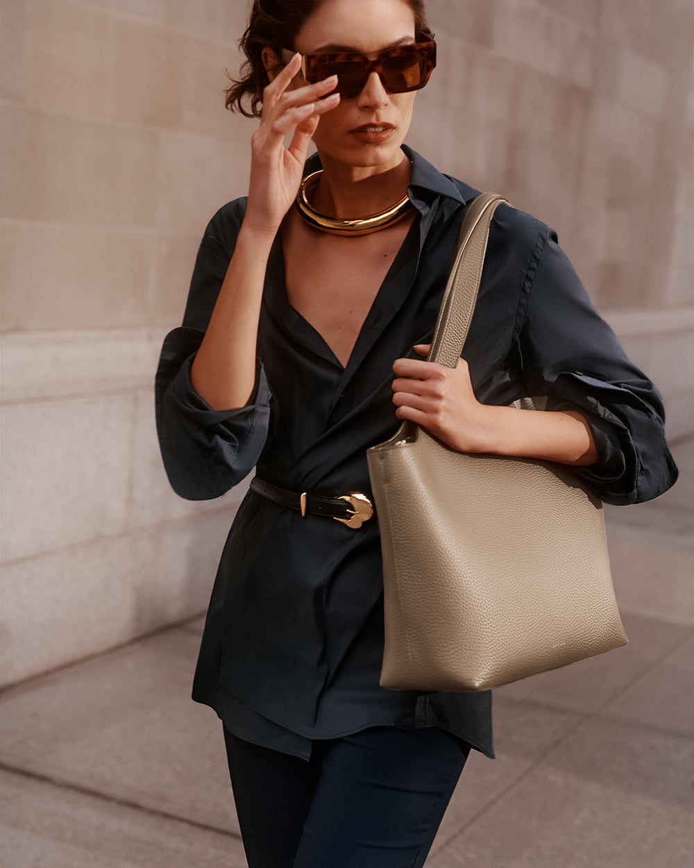 Woman wearing sunglasses, a necklace, and a belted top, holding a shoulder bag.