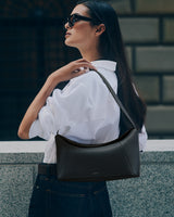 Person wearing sunglasses and holding a handbag over shoulder, standing outdoors.