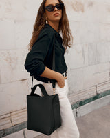 Person wearing sunglasses with a shoulder bag, blouse, and pants, standing against a stone wall