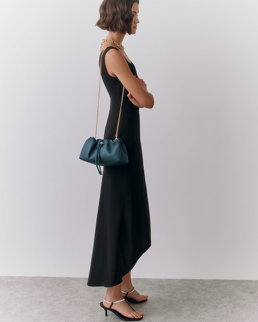 Woman in long dress with shoulder bag and sandals, standing against plain backdrop.