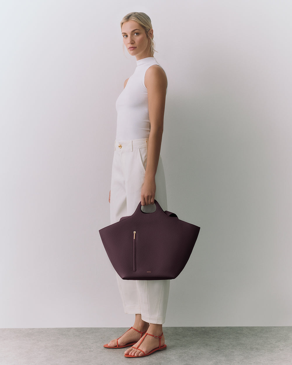 Person standing, holding a large handbag, wearing a sleeveless top and pants with sandals.