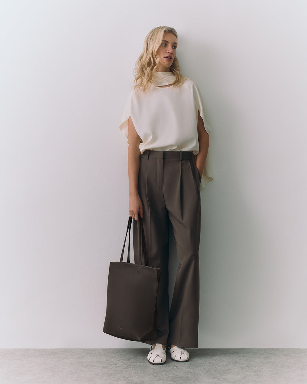 Woman in sleeveless top and trousers holding a tote bag, standing against a plain wall.