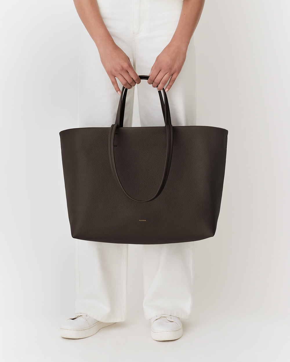 Person holding large tote bag by handles, wearing pants and shoes.