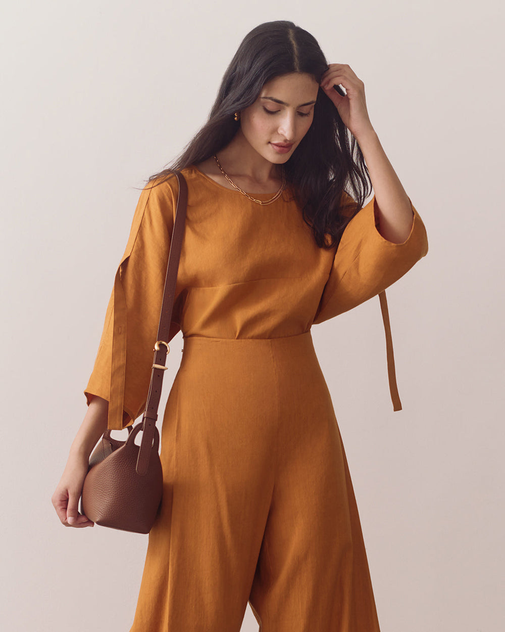 Person wearing a jumpsuit with a crossbody bag, looking down, touching hair.