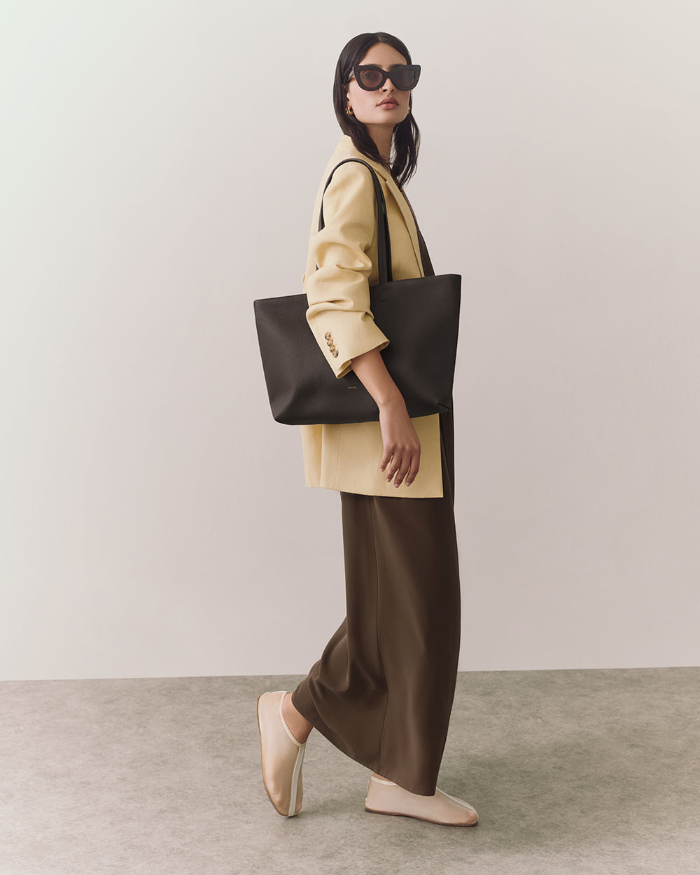 Person wearing sunglasses, a jacket, long pants, and carrying a large tote bag