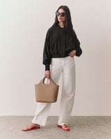 Person wearing sunglasses, jacket, pants, sandals, holding a handbag, stands indoors.
