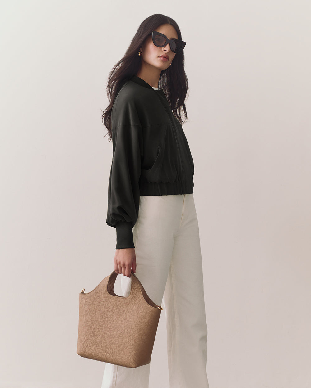 Woman in jacket and pants holding a handbag, wearing sunglasses.