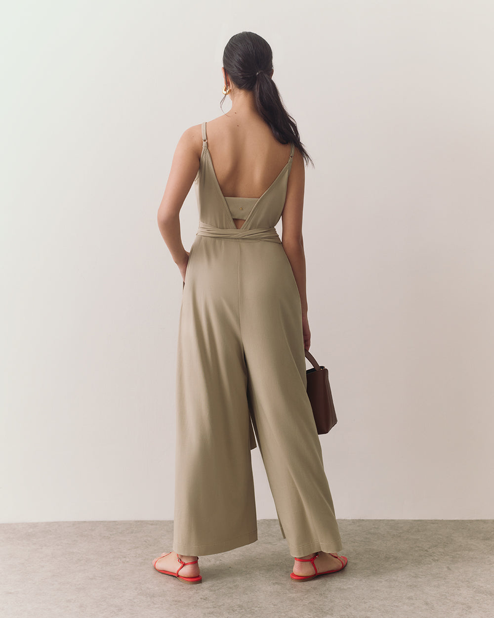 Person wearing a sleeveless jumpsuit and sandals, holding a handbag, standing.