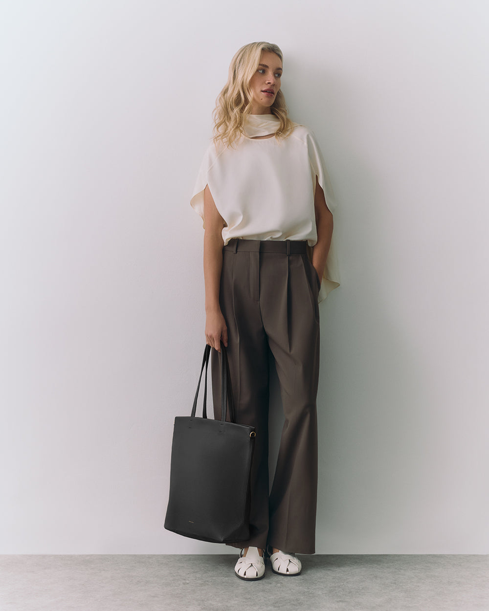 Person stands against wall holding tote bag, wearing loose top and trousers.