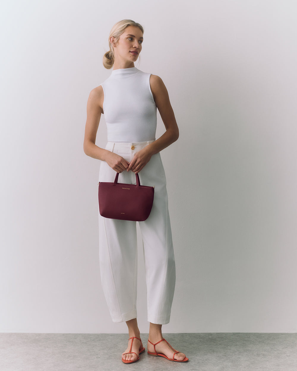 Person in sleeveless top and loose pants holding a handbag, wearing sandals, standing indoors.