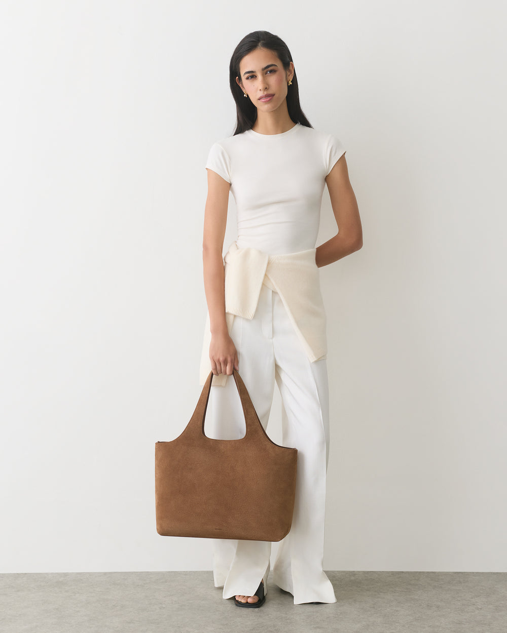 Woman in a top and pants holding a large suede bag, with a sweater tied at the waist.