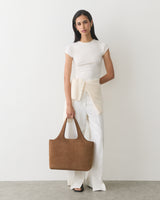Woman in a top and pants holding a large suede bag, with a sweater tied at the waist.