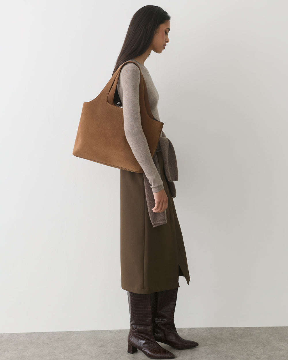 Woman in long skirt and boots with large suede shoulder bag standing sideways.
