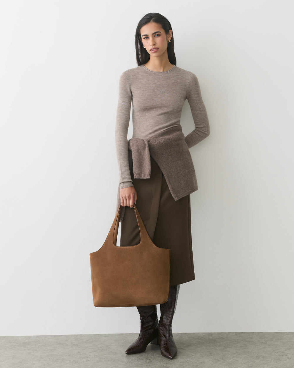 Person holding suede large bag, wearing sweater and skirt, standing against plain wall.