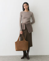 Person holding suede large bag, wearing sweater and skirt, standing against plain wall.
