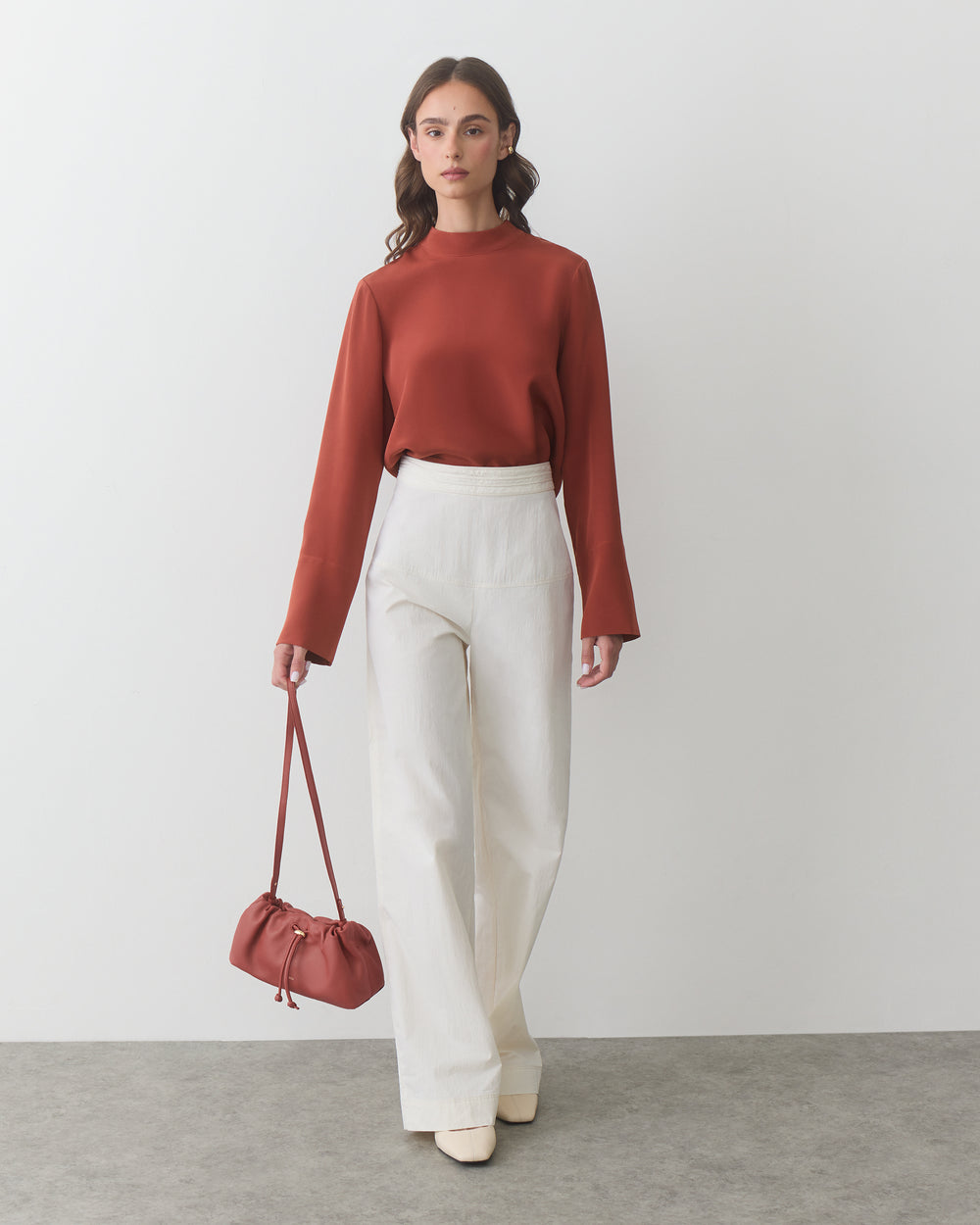 A person stands wearing a long-sleeve top and trousers, holding a small bag.