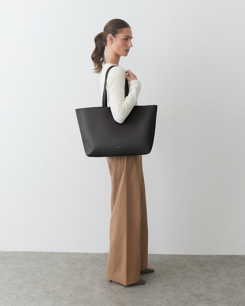 Person standing with large tote bag, wearing long-sleeve top and wide-leg pants.