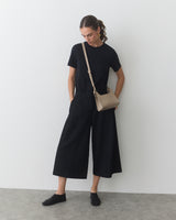 Person in wide-leg pants and shirt, with a crossbody bag, standing against a plain wall.