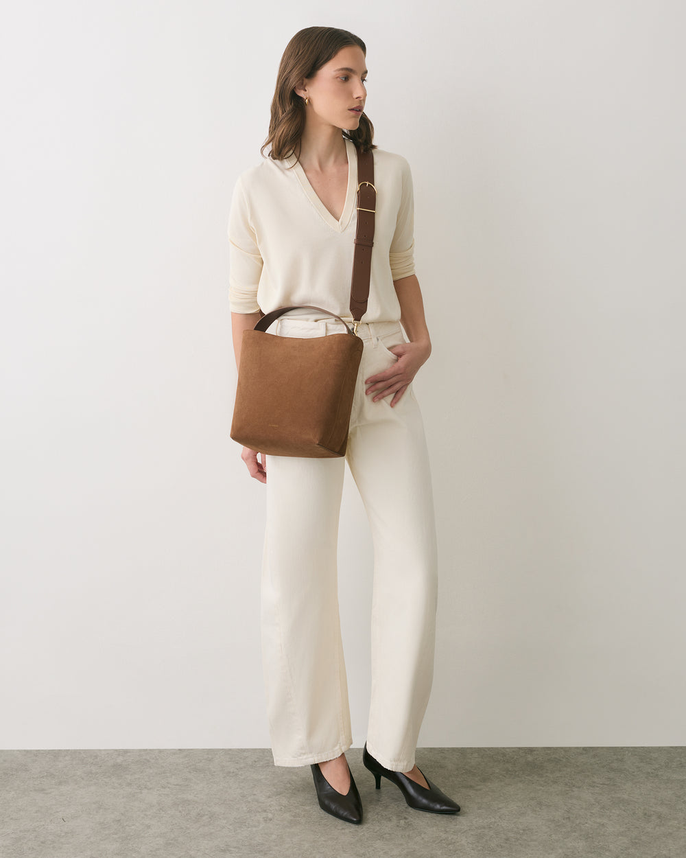 Person in casual outfit holding a shoulder bag wearing heels standing against a plain backdrop.