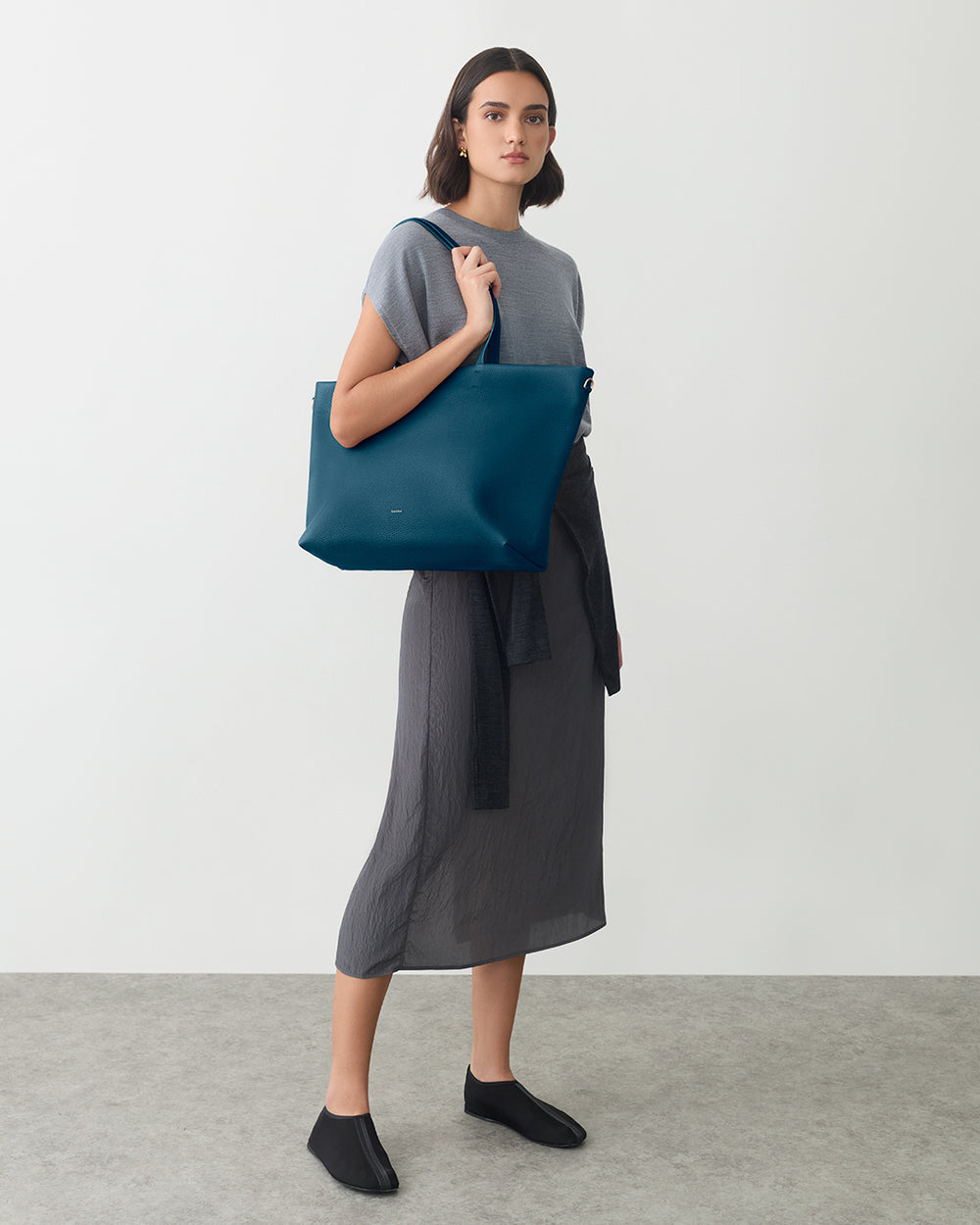 Person holding tote bag, wearing dress and shoes, standing on indoor floor.