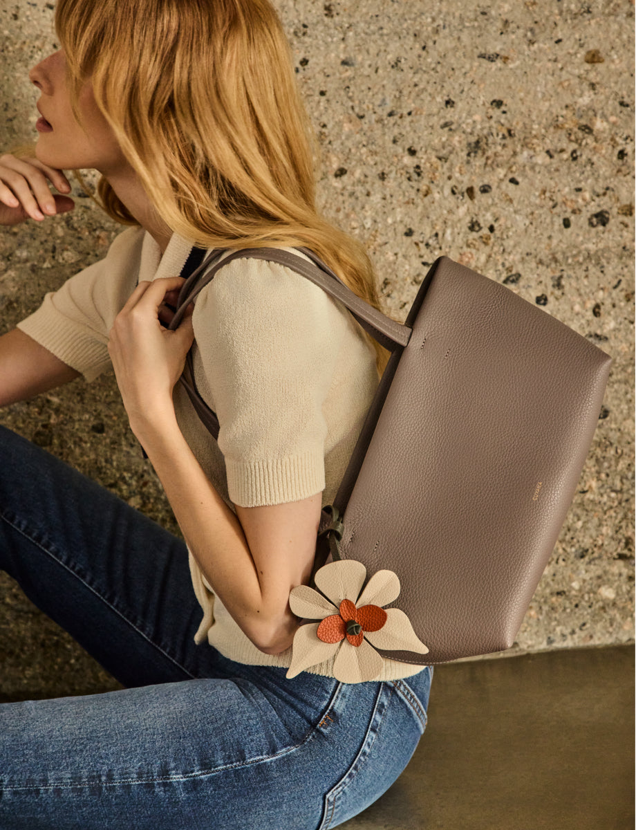 Person holding a handbag with a flower charm, sitting against a textured wall.