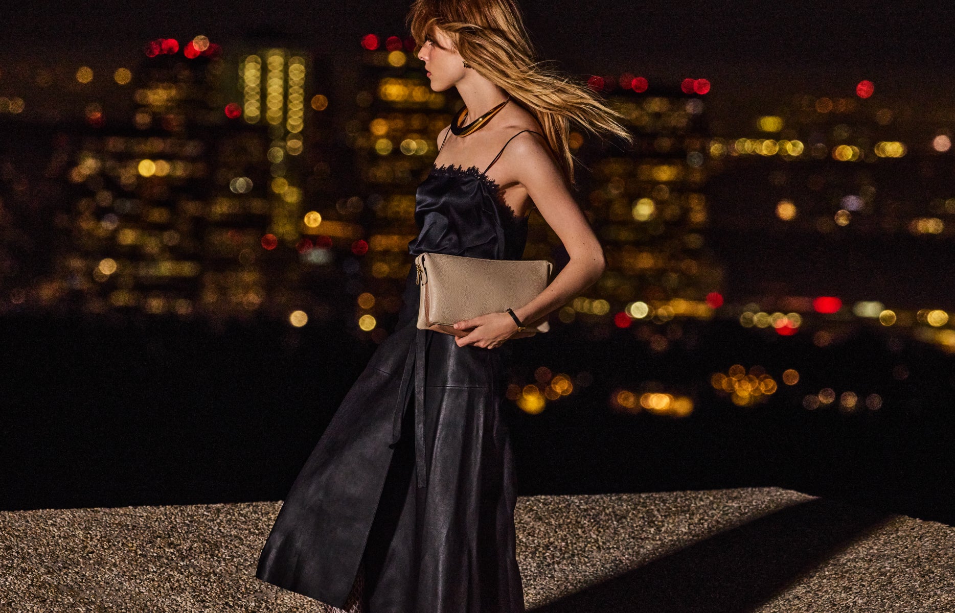 Person walking on rooftop at night with city lights in background, holding a clutch bag.