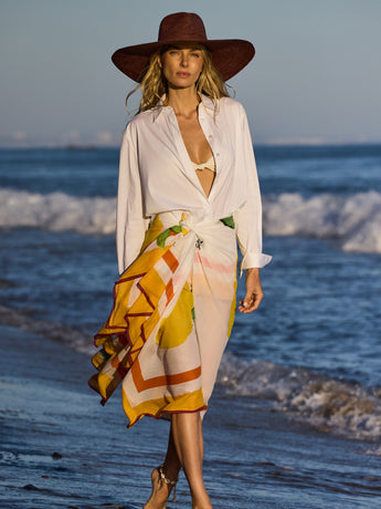 Person on beach wearing wide-brim hat, open shirt, and patterned wrap skirt.