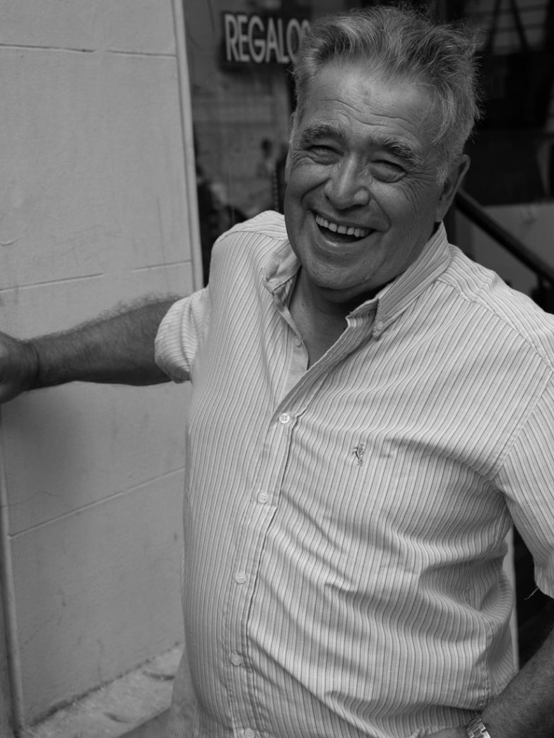 Older man smiling and leaning against a wall.