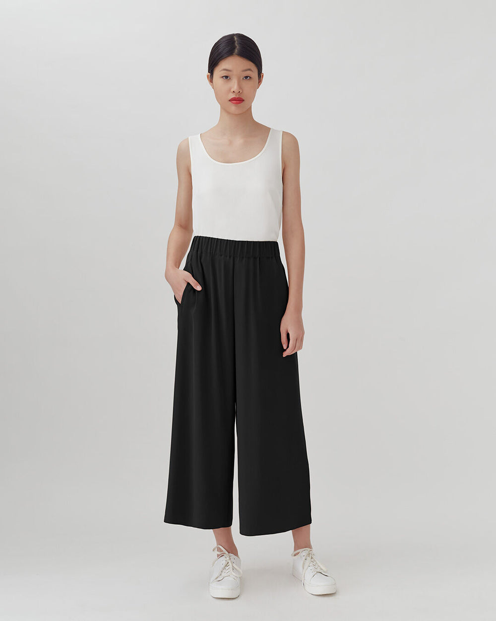 Woman standing in sleeveless top and loose pants, wearing sneakers.