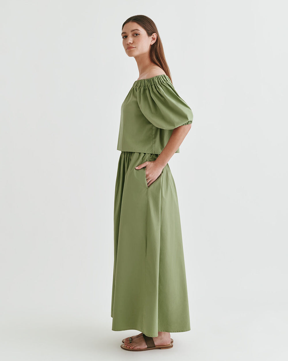 Woman standing in a studio with hands on hips, wearing a long dress and sandals.