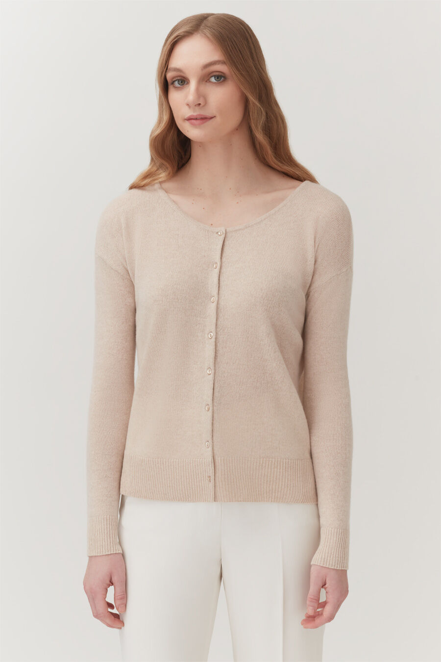Woman in a buttoned sweater and pants standing against a plain backdrop.