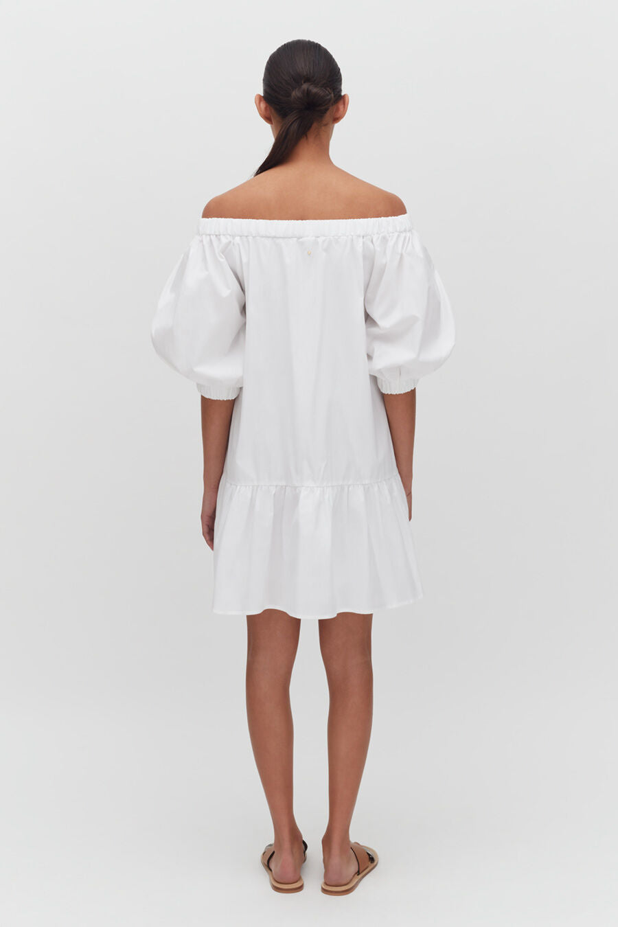 Woman standing with back facing camera, wearing a knee-length dress and sandals.